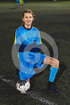 little girl in a blue uniform kneeling in a field and taking a photo with a soccer ball, vertical shot