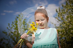 A little girl with a bouquet of yellow dandelion flowers and green trees with a blue sky in the background. Portrait of