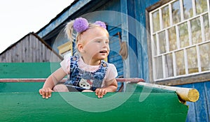 A little girl with bows looking surprisingly sitting on a cart on the background of a village house