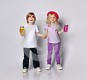 Little girl and boy in colorful casual clothes. Smiling, holding hands, yellow and pink cocktail bottles. Posing isolated on white