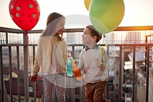 Little girl and boy holding balloons while enjoying a drink on the terrace of an apartment. Family, together, party