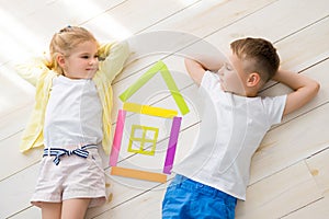 A little girl with a boy lie on the floor next to a house of colored paper. View from above