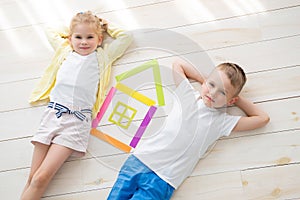 A little girl with a boy lie on the floor next to a house of colored paper. View from above