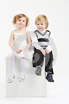 Little girl and boy sit on white big cube on white
