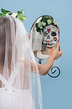 Little girl in bridal gown wears painted scary skull mask looking through mirror