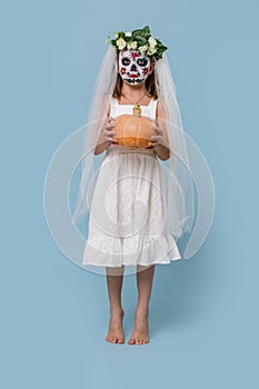 Little girl in bridal gown wears painted scary skull mask over blue background