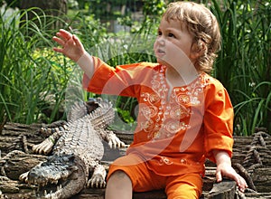 A little girl in bright orange clothes is sitting next to a stuffed crocodile on a background of green grass