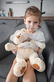 Little girl with broken finger holds teddy bear with a bandaged paw at the doctor& x27;s appointment in the hospital