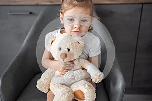 Little girl with broken finger holds teddy bear with a bandaged paw at the doctor's appointment in the hospital