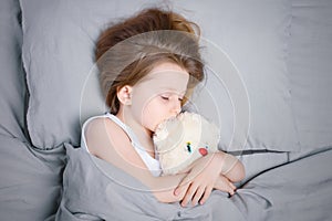 A little girl with brown hair is sleeping on her side with a stuffed toy in the bed under the blanket. Solid gray bed linen top