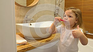 A little girl brushes her teeth in the bathroom and gives a thumbs up