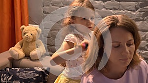 Little girl brushing her mother`s hair with hairbrush, modern living room background, family concept indoors