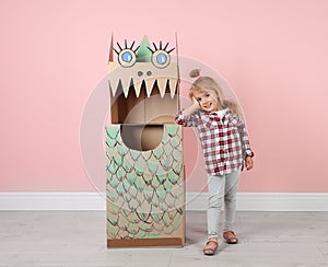 Little girl and cardboard costume of dinosaur near color wall indoors.