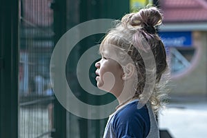 A little girl carefully looks over the fence at the children`s playground. Concept: the desire to play sports, the limitations of