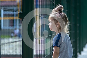 A little girl carefully looks over the fence at the children`s playground. Concept: the desire to play sports, the limitations of