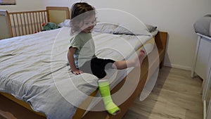 A little girl with a cast on her leg is sitting on a bed in a room.