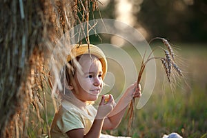 A little girl of Caucasian appearance with light hair in a field with stacks and ears of wheat.