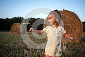 A little girl of Caucasian appearance with light hair in a field with stacks and ears of wheat.