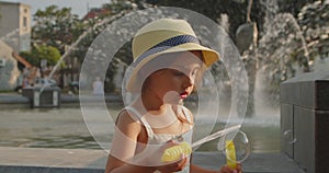 Little girl cheerfully blows soap bubbles on the background of a fountain.