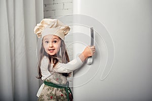 Little girl in a chef hat looks into the refrigerator