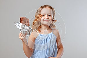 Little girl child blonde holds a bitten bar of chocolate in her hands. Gray background, studio, portrait