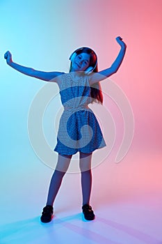 Little girl, child in headphones dancing, listening to music in headphones over gradient blue pink studio background in