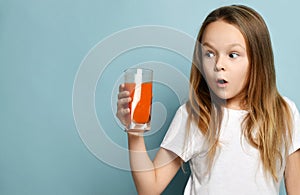 Little girl child kid drinking carrot orange juice surprised in white copy space blank t-shirt on light blue background