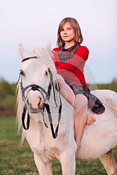 Little girl child in a red dress sits astride a white horse