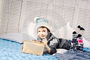 Little girl the child sitting in pajamas and hat on the bed with garland of light bulbs with gifts boxes wrapped in a non-colored