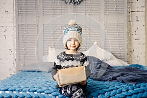 Little girl the child sitting in pajamas and hat on the bed with garland of light bulbs with gifts boxes wrapped in a non-colored photo