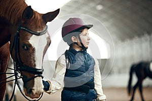 Little girl, child in special uniform and helmet walking with horse during educational course of horseback riding