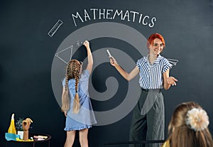 Little girl, child standing at chalkboard, drawing triangle, doing math tasks with teacher. Learning geometry at school.