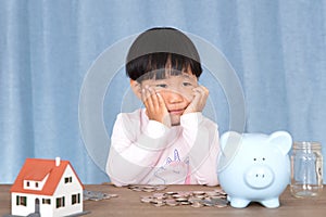 Little girl with chin resting on both hands is meditating in front of a pile of dollar coins and piggy bank