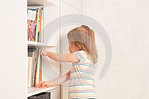 Little girl chooses a book in a bookcase at home.