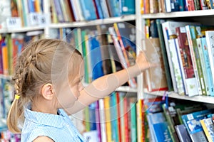 Little girl chooses, takes book with fairy tales from shelf in children`s library.Special reading kids room.Many shelves with