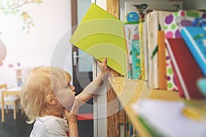 Little girl choosing and taking book from shelf to read