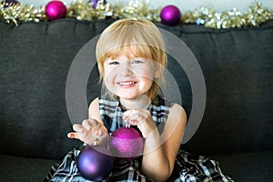 Little girl with Christmas balls sitting on dark grey couch, smiling and happy