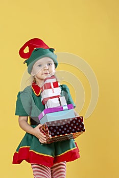Little girl in a Christmas elf costume, carries a stack of gifts. Photo on a yellow background.