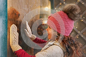 Little girl on christmas evening looks into the window showcase with gifts in anticipation of a miracle