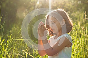 Little Girl closed her eyes, praying in a field during beautiful sunset. Hands folded in prayer concept for faith