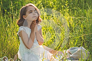 Little Girl closed her eyes, praying in a field during beautiful sunset. Hands folded in prayer concept for faith