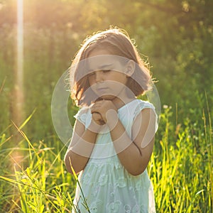 Little Girl closed her eyes, praying in a field during beautiful sunset. Hands folded in prayer concept for faith