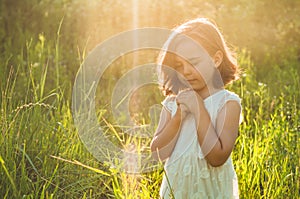 Little Girl closed her eyes, praying in a field during beautiful sunset. Hands folded in prayer concept for faith