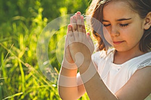 Little Girl closed her eyes, praying in a field during beautiful sunset. Hands folded in prayer concept for faith