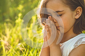 Little Girl closed her eyes, praying in a field during beautiful sunset. Hands folded in prayer concept for faith