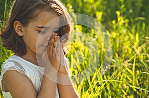 Little Girl closed her eyes, praying in a field during beautiful sunset. Hands folded in prayer concept for faith