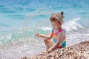 A little girl in a colorful swimsuit sits by the sea. Girl is playing on the beach. Beautiful and bright blue sea