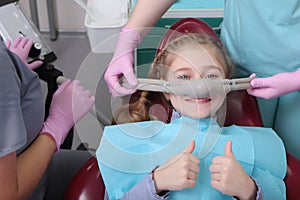 A little girl is comfortable to treat her teeth under superficial sedation. The girl smiles and holds two thumbs up. Milk teeth