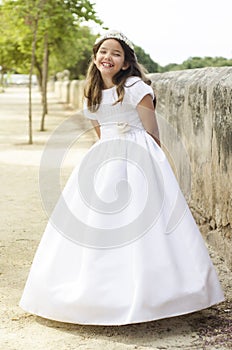 Little girl in communion dress smiles with her arms crossed behind. Full body photography of the dress, sunbath