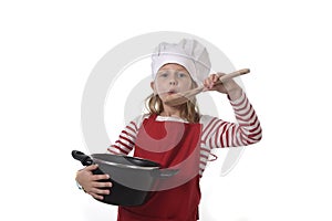 Little girl in cooking hat and red apron playing cook smiling happy holding pot and pretending tasting food